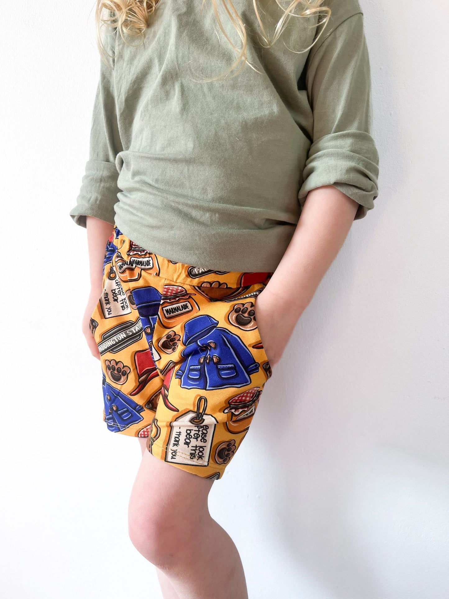 Child wearing British Bear -themed jogger shorts with golden-yellow background, blue coats, marmalade jars, and pockets.