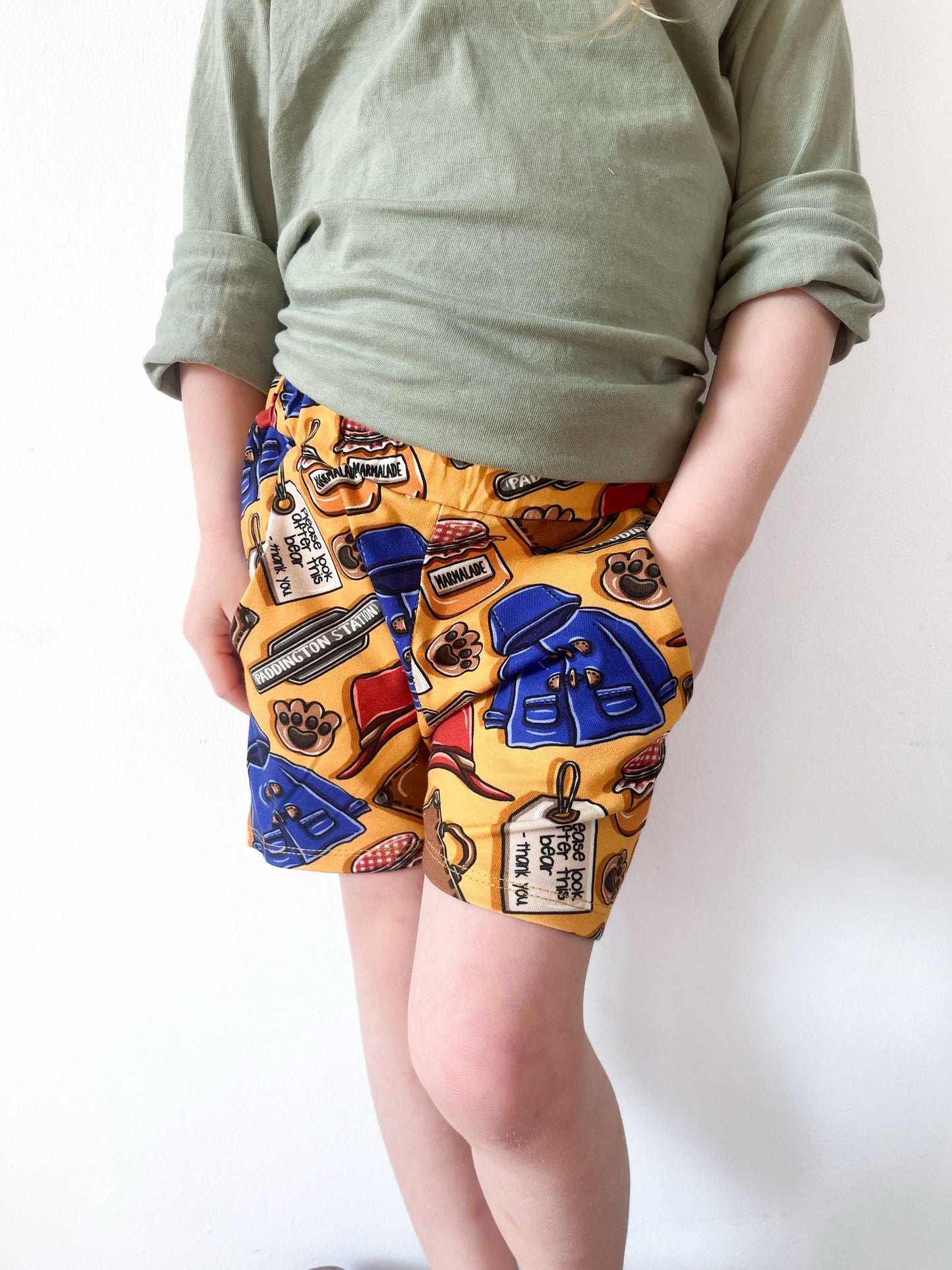 Child wearing British Bear -themed jogger shorts with golden-yellow background, blue coats, marmalade jars, and pockets.